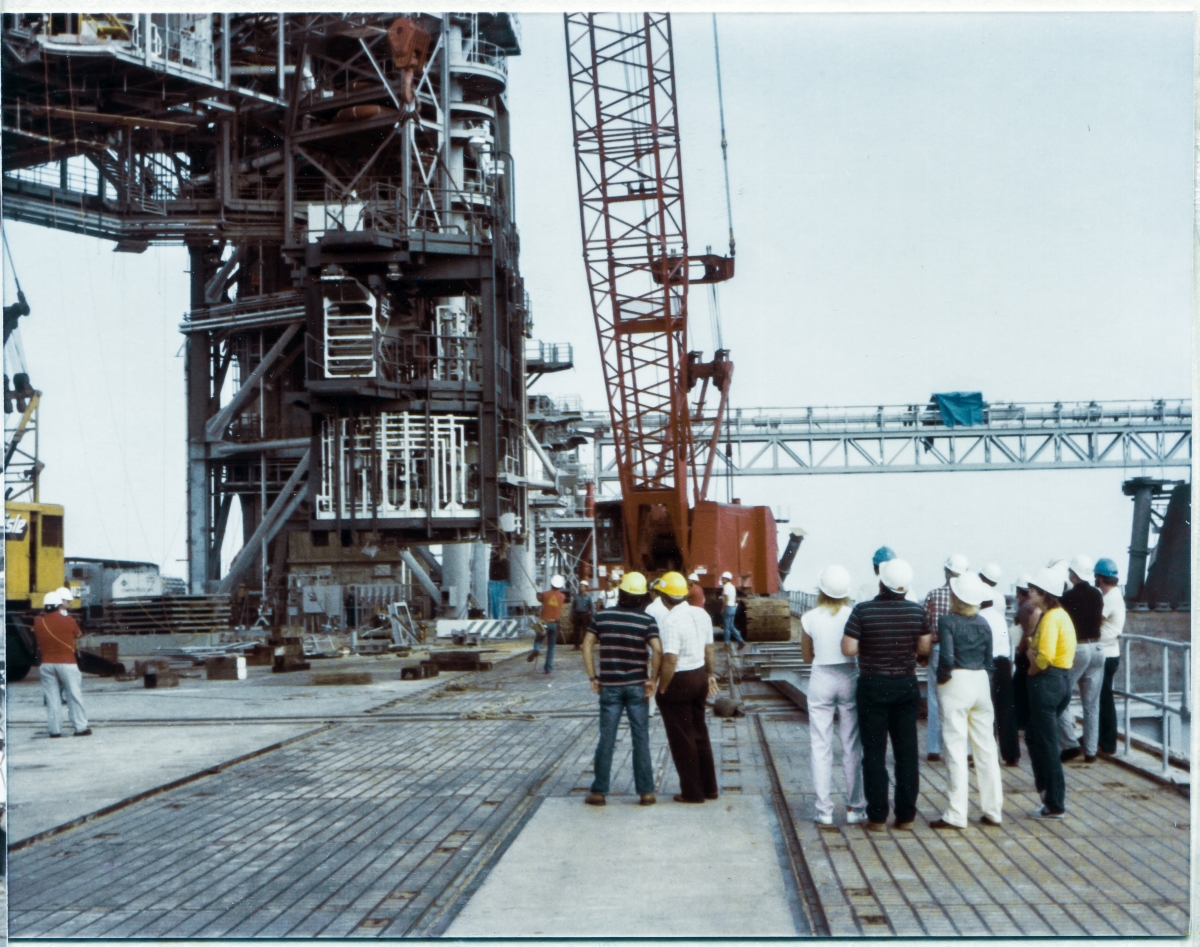 Image 089. At Space Shuttle Launch Complex 39-B, Kennedy Space Center, Florida, the OMBUU begins its vertical rise, up and away from the Pad Deck, in earnest. The big red Manitowoc crawler-crane which is lifting it can now be seen clearly. Seen in the distance, wearing a white shirt and white hardhat which contrasts well against the darker background of the crane, wearing blue jeans, Wade Ivey, owner of Ivey Steel Erectors, has entered the dynamic of the Lift as a participant, keeping a close eye on everything, as Union Ironworkers from Local 808, both wearing red shirts, haul on their respective tag lines that are attached to the bottom corners of the OMBUU, keeping it aligned properly as it heads upward away from them. In the near foreground, personnel from various construction contractors and oversight agencies witness the lift, in progress, from a safe-enough distance. Photo by James MacLaren.